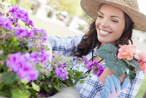 A woman in a garden wearing a wide-brimmed straw hat, smiling as she tends to a variety of flowering plants in a neatly arranged outdoor space. The garden features a well-maintained lawn with dense, green grass, bordered by flower beds containing vibrant purple, pink, and orange blooms. In the background, there are several mature trees and neatly trimmed hedges, suggesting a landscaped backyard or front garden in Wanstead. She is dressed in a blue plaid shirt and gardening gloves, indicating active gardening work. The scene is lit by natural daylight, with a bright, cheerful atmosphere that highlights the diversity of plants and the tidy layout of the garden, reflecting professional gardening and lawn care services provided by Lawn Mowing Wanstead in the local area.
