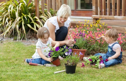 A woman with short blonde hair dressed in a white blouse and jeans is sitting on a well-maintained, lush green lawn in a garden in Wanstead, engaging with two young children. The children, a girl and a boy, are seated on the grass and are actively involved in planting and tending to vibrant flower pots filled with purple, pink, and yellow blooms. The garden features a background of various plants including a dense cluster of ornamental grasses or shrubs near a wooden deck or patio, with a brick or wooden structure visible behind. The scene is illuminated by natural daylight, suggesting a pleasant, mild weather condition suitable for outdoor gardening activities. The setting emphasizes the tidy, landscaped outdoor space typical of Wanstead gardens, showcasing a combination of lawn, flower beds, and garden planting activities, aligning with the professional outdoor maintenance and gardening services provided by Lawn Mowing Wanstead.